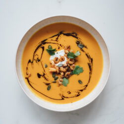 Kürbissuppe mit Ingwer und Curry in heller Schale, getoppt mit gebratenen Garnelen, Sahne, Kürbiskernöl, Kürbiskernen und frischem Koriander