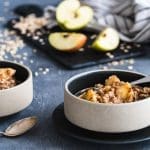 Zwei Schalen Porridge mit Apfel und Zimt, dahinter Apfelhälften und Haferflocken