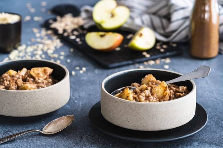 Zwei Schalen Porridge mit Apfel und Zimt, dahinter Apfelhälften und Haferflocken