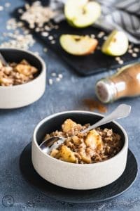 Porridge mit Apfel und Zimt in Schalen, daneben Äpfel und Haferflocken