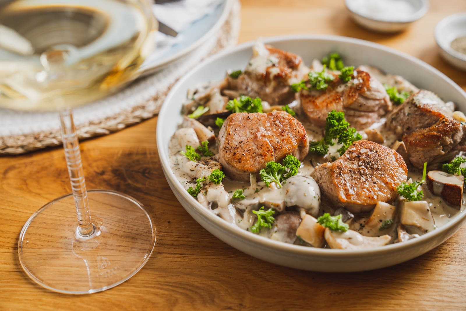 Schweinemedaillons in Champignonrahmsoße in einer Schüssel, daneben ein Glas Riesling