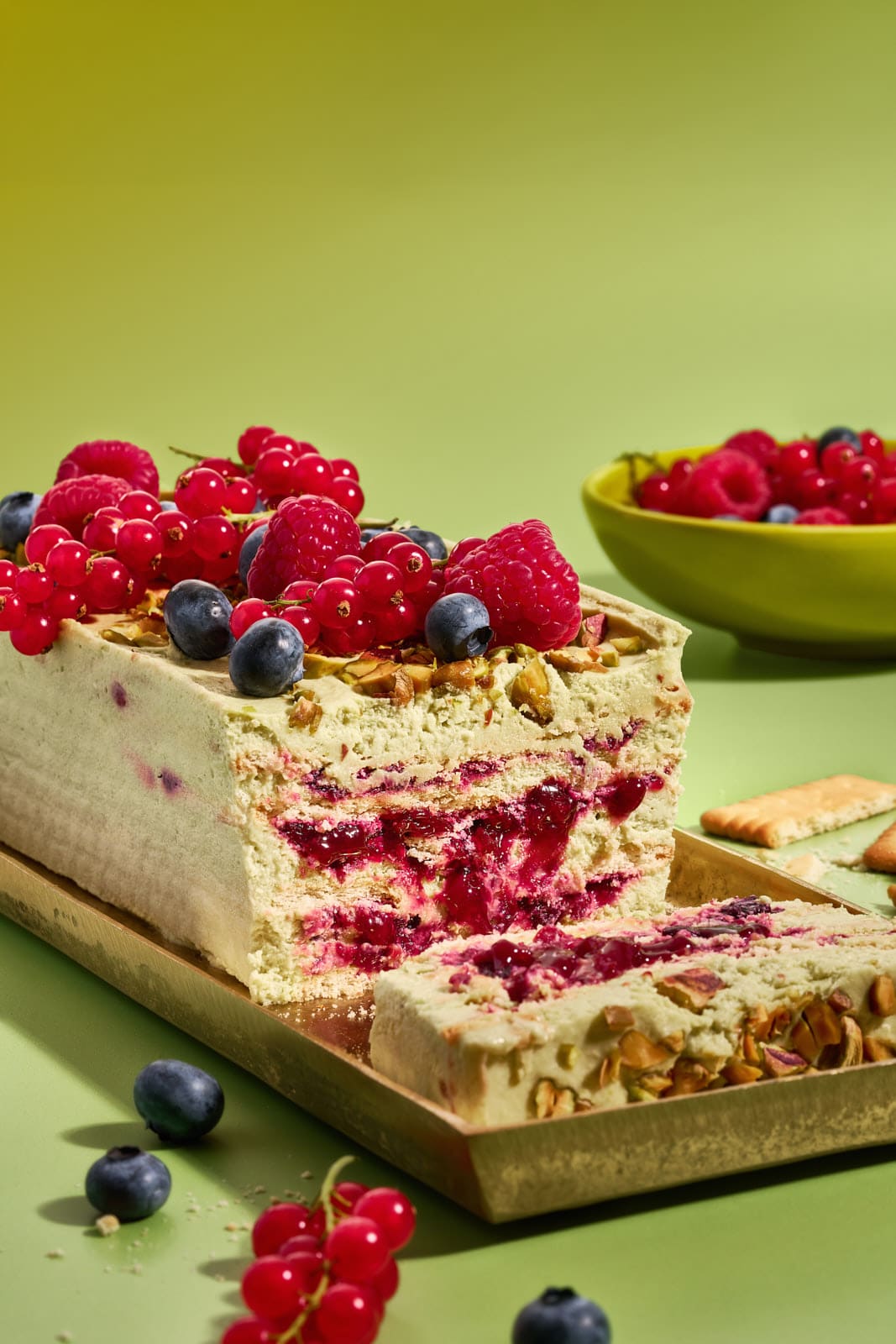 Angeschnittener Pistazienkuchen ohne Backen, Schnittfläche mit Pistaziencreme-Schichten und Waldbeeren-Kompott, oben frische Beeren