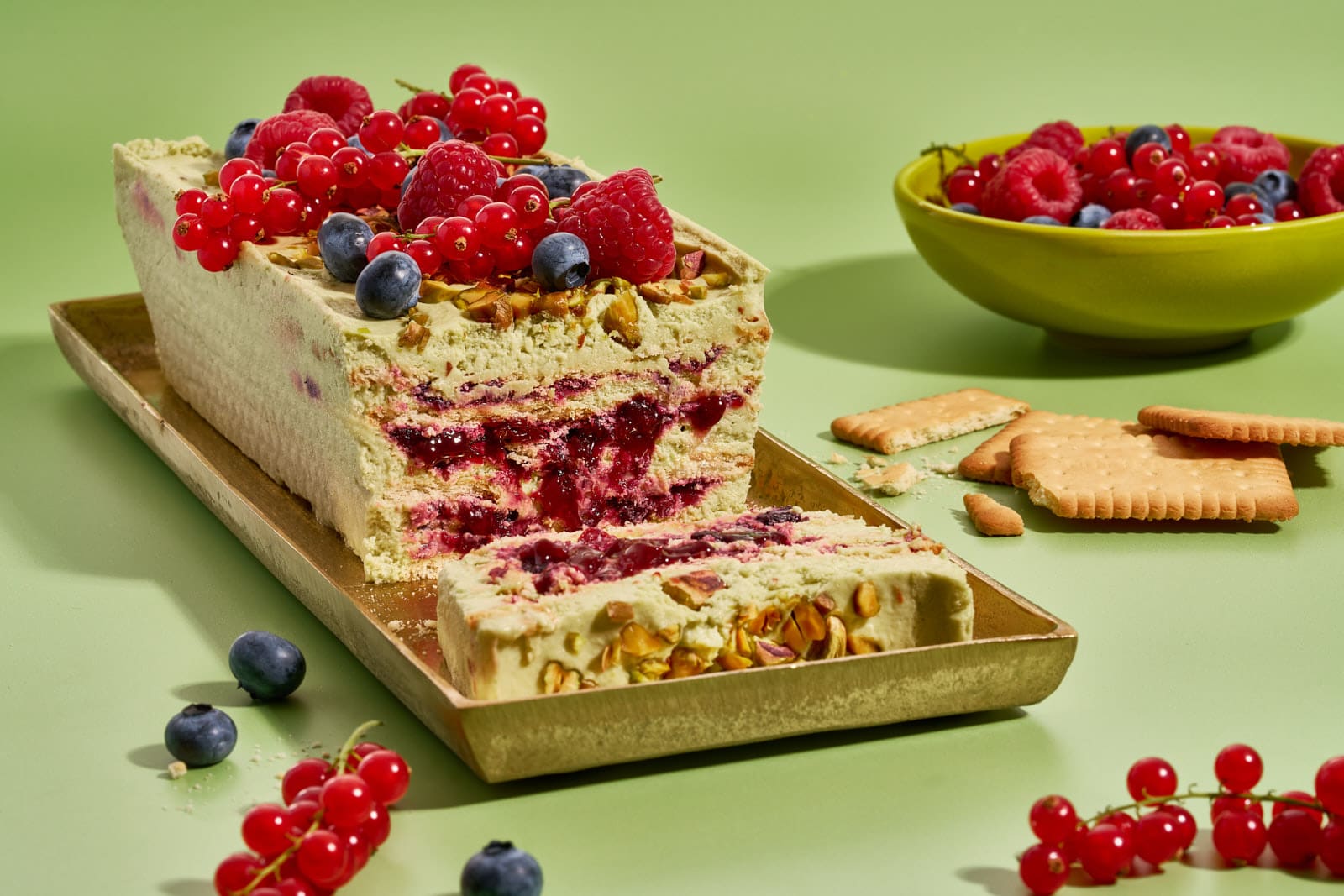 Pistazienkuchen ohne Backen mit Waldbeeren auf goldenem Tablett, angeschnitten, daneben Butterkekse und eine Schale frische Beeren