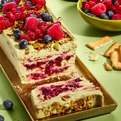 Pistazienkuchen ohne Backen mit Waldbeeren auf goldenem Tablett, daneben Butterkekse und eine Schale mit frischen Beeren