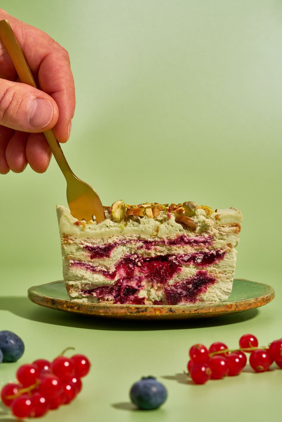 Goldene Kuchengabel sticht in ein Stück Pistazienkuchen ohne Backen mit Waldbeeren auf grünem Teller