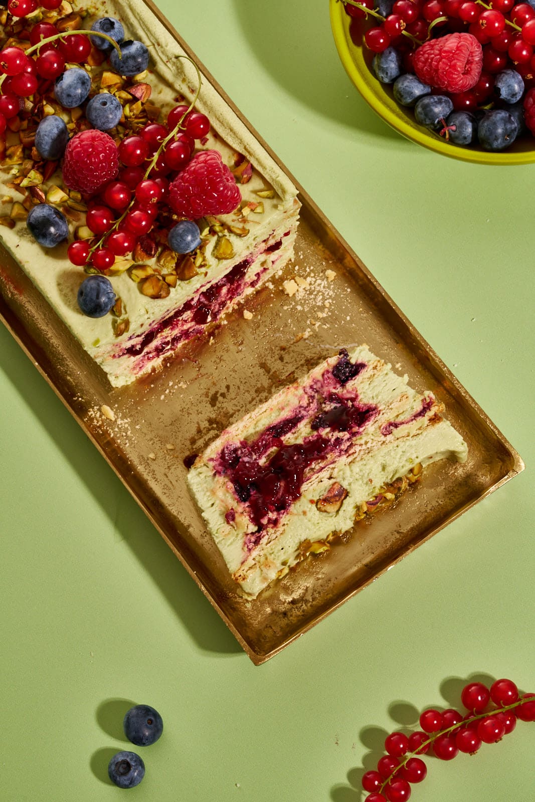 Pistazienkuchen ohne Backen auf goldenem Tablett, seitliche Ansicht mit sichtbarem Waldbeeren-Kompott zwischen den Cremeschichten