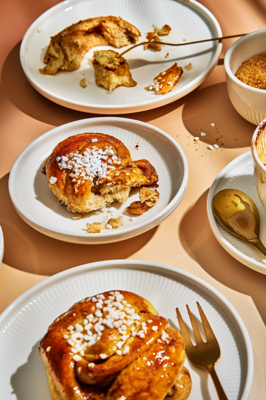 Kanelsnurrer. Dänische Zimtknoten mit Kardamom 3 Mehrere Teller mit gedrehten, karamellisierten Zimtknoten mit Hagelzucker, teils angebissen, dazu Kaffee und goldene Löffel