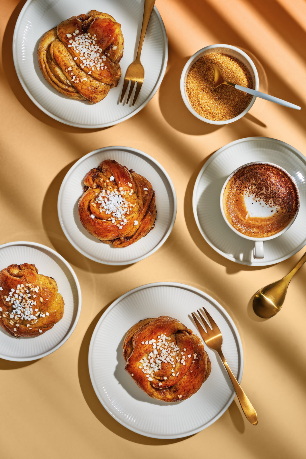 Kanelsnurrer. Dänische Zimtknoten mit Kardamom 1 Goldbraune Kanelsnurrer mit Hagelzucker auf Tellern, daneben Zuckerschale und Cappuccino auf sandfarbenem Hintergrund
