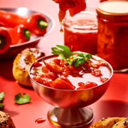 Ein Stück eingelegte Paprika auf einer Gabel über einer Schale Antipasti, im Hintergrund gefüllte Gläser