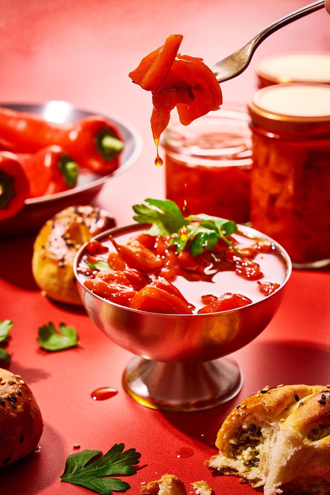 Ein Stück eingelegte Paprika auf einer Gabel über einer Schale Antipasti, im Hintergrund gefüllte Gläser