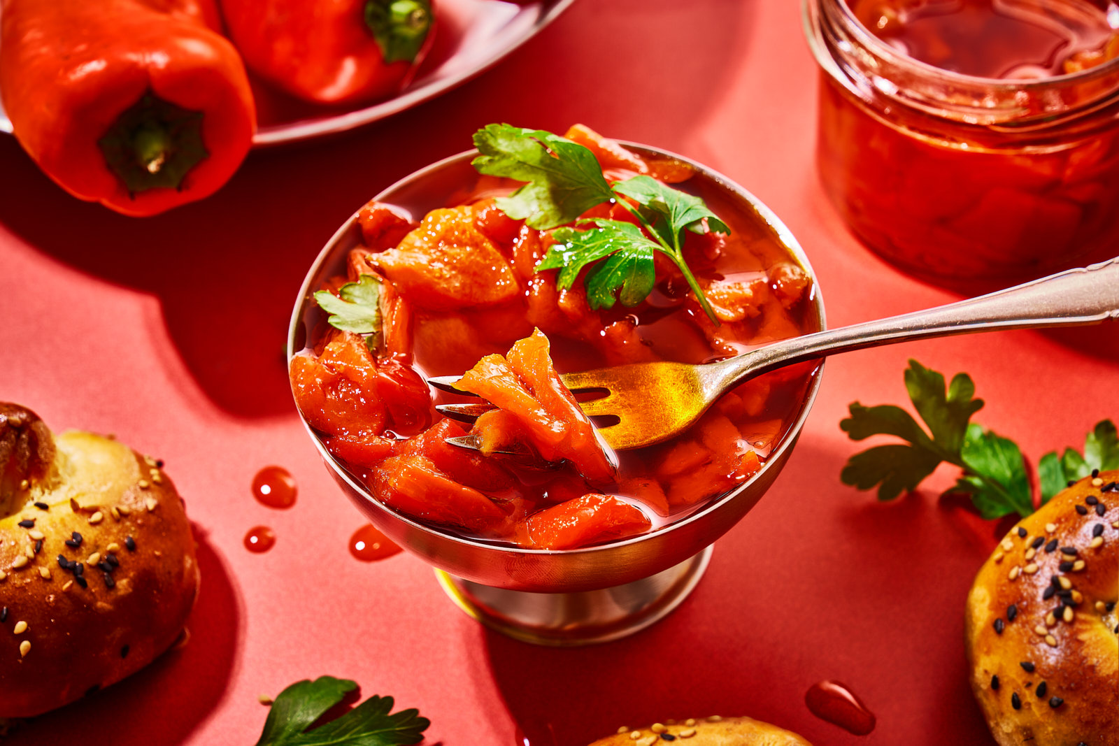 Eingelegte Paprika mit Gabel in einer Schale, daneben frische rote Paprikaschoten und Brötchen