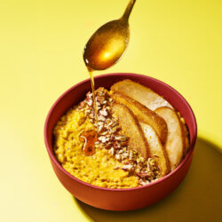 Pumpkin Spice Porridge in roter Schale mit karamellisierten Birnenspalten, Pekannüssen und Ahornsirup vom Löffel